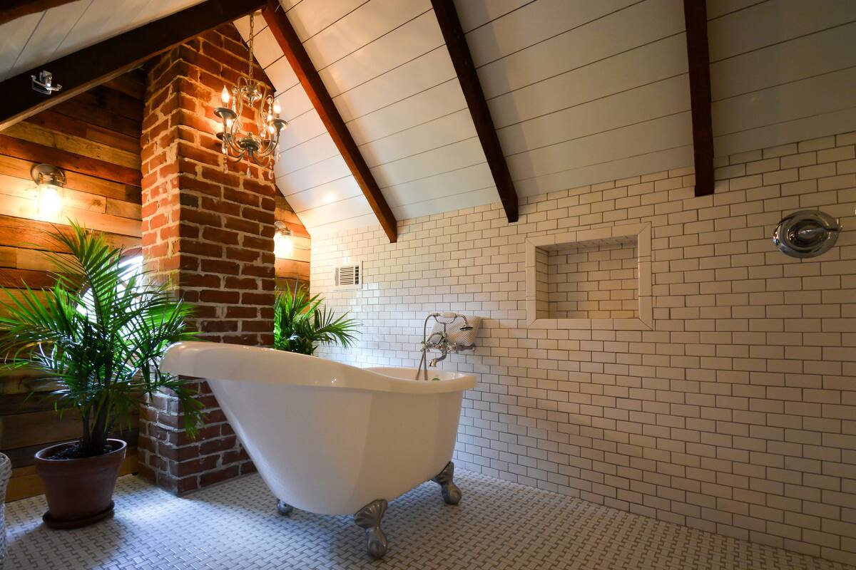 Une salle de bain avec une baignoire sur pieds, des murs recouverts de carreaux blancs, une colonne en briques et des poutres en bois au plafond.