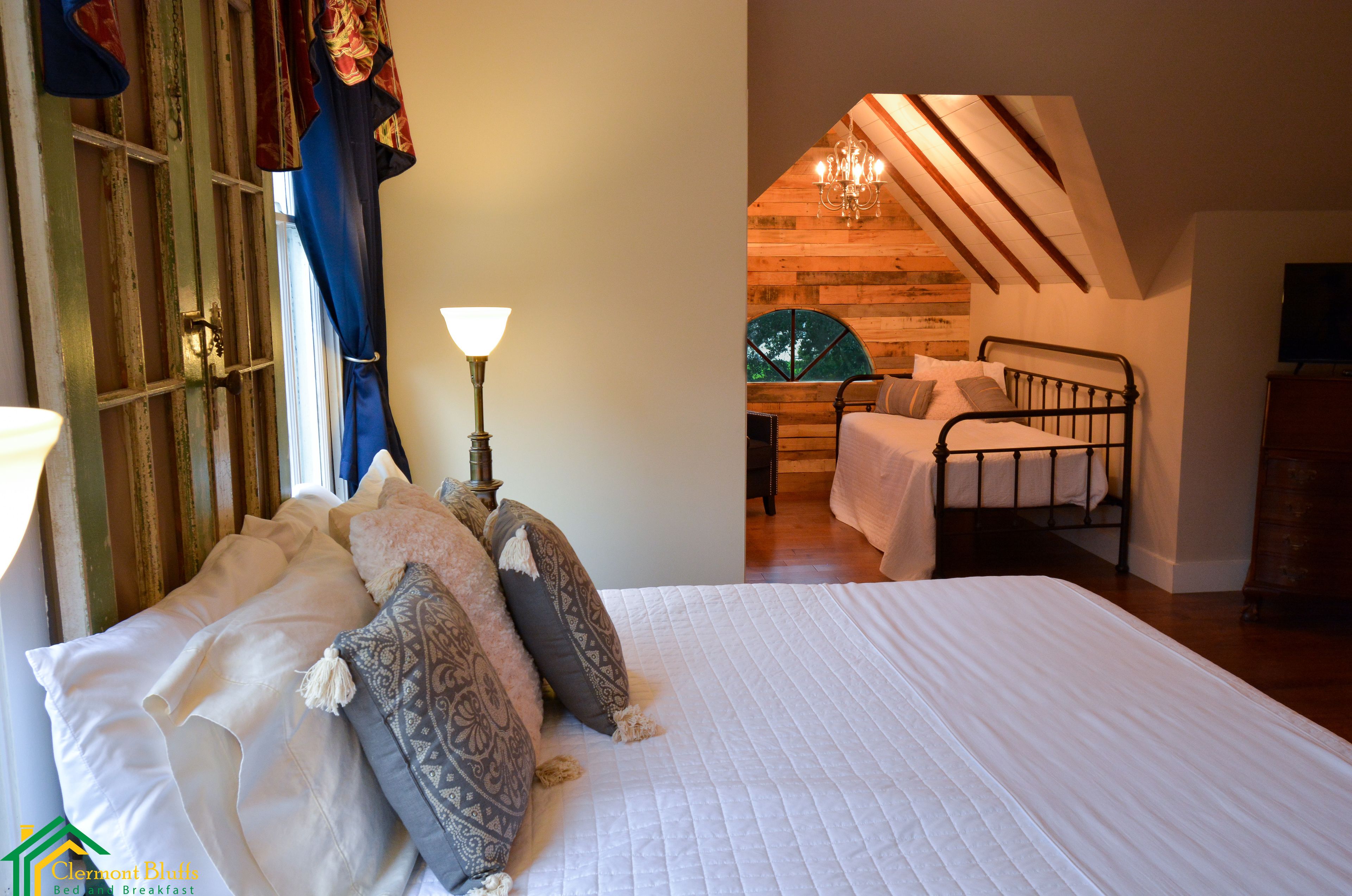 A bedroom with a large bed, a nightstand, a lamp, and a view of a dining area with a chandelier.
