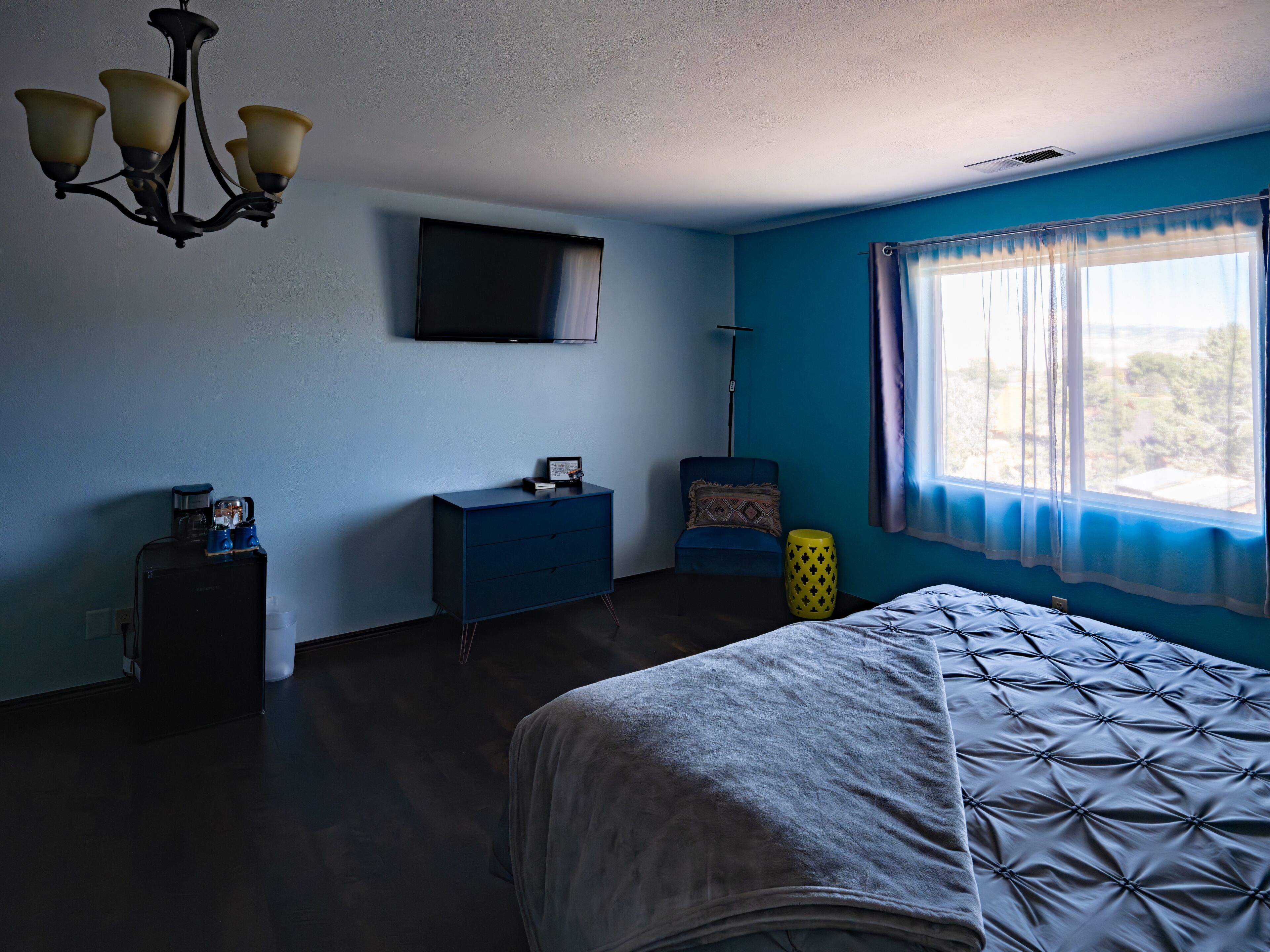 Un dormitorio con una cama, un aparador, un televisor en la pared, una silla y una ventana con cortinas.