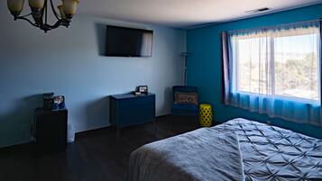 Un dormitorio con una cama, un aparador, un televisor en la pared, una silla y una ventana con cortinas.