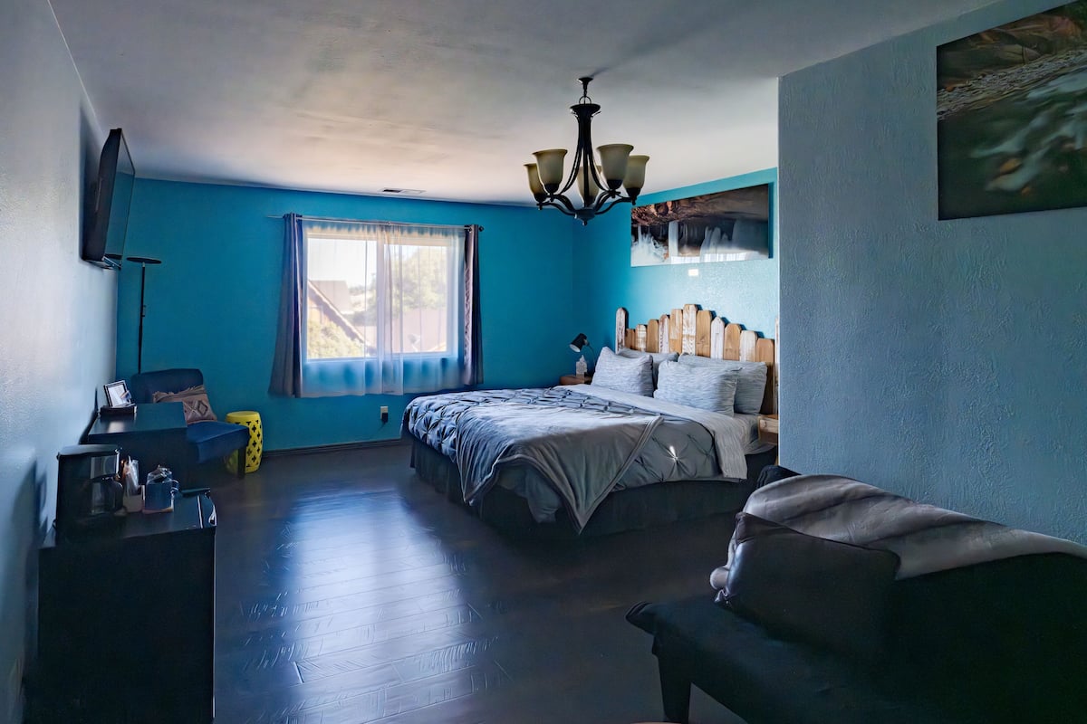 Un dormitorio con cama, una silla, un escritorio, una ventana con cortinas y un cuadro en la pared.