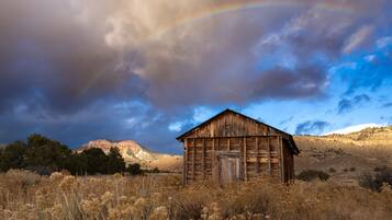 A rustic wooden barn in a dry field with a mountain backdrop and a rainbow.