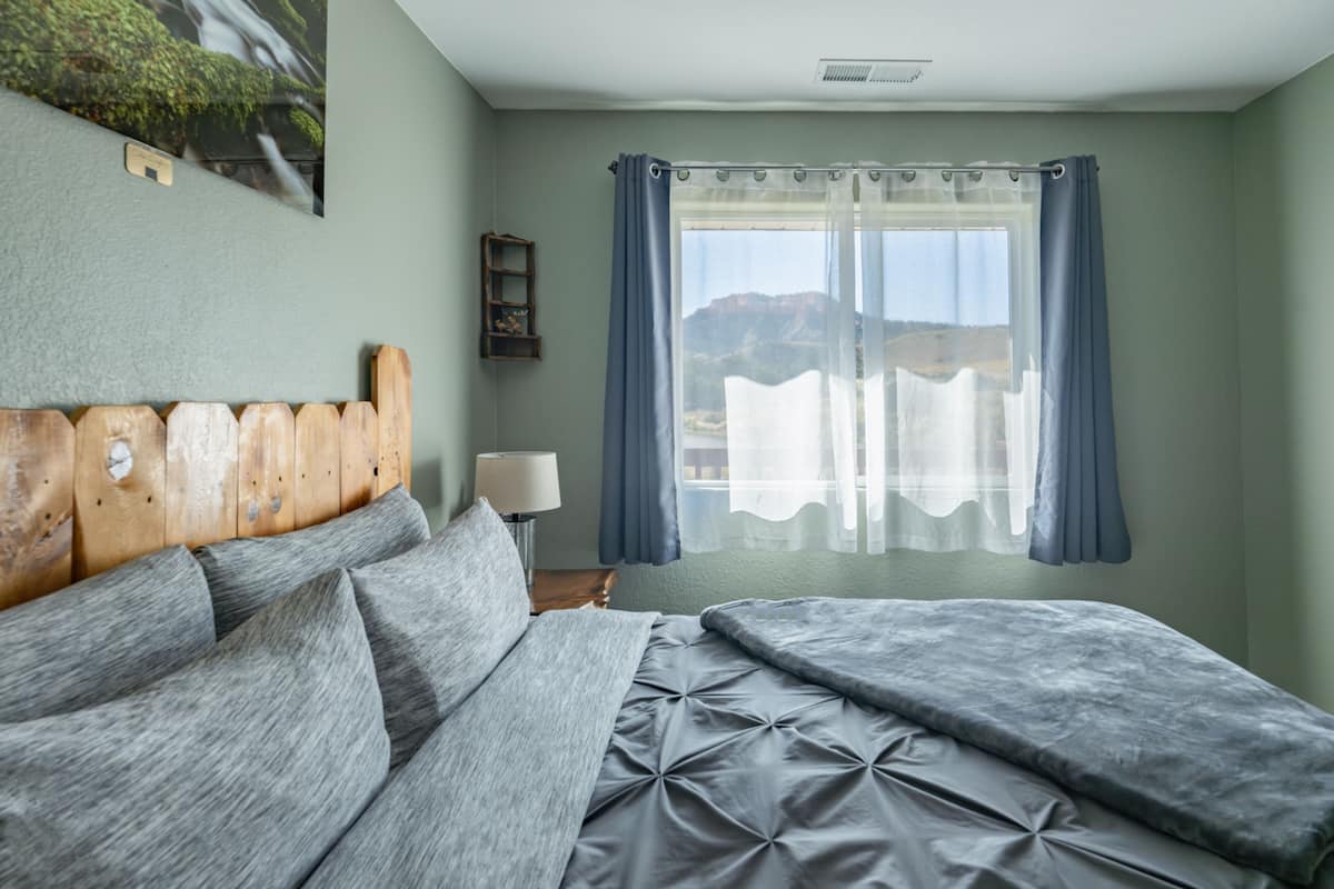 Un dormitorio con cabecera de madera, cama con ropa de cama gris, lámpara de noche y una ventana con cortinas azules.