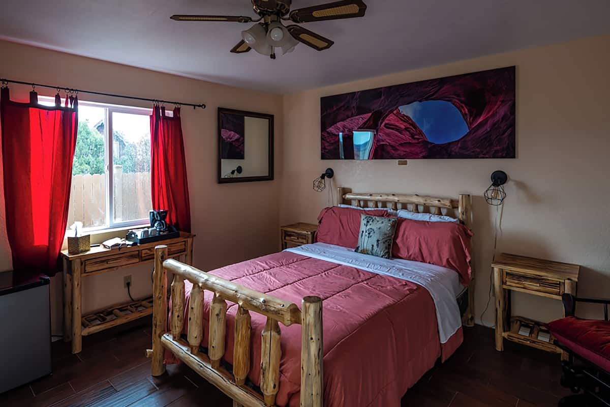 Un dormitorio con cama de madera, cortinas rojas, ventilador de techo, un cuadro en la pared y un escritorio con silla.