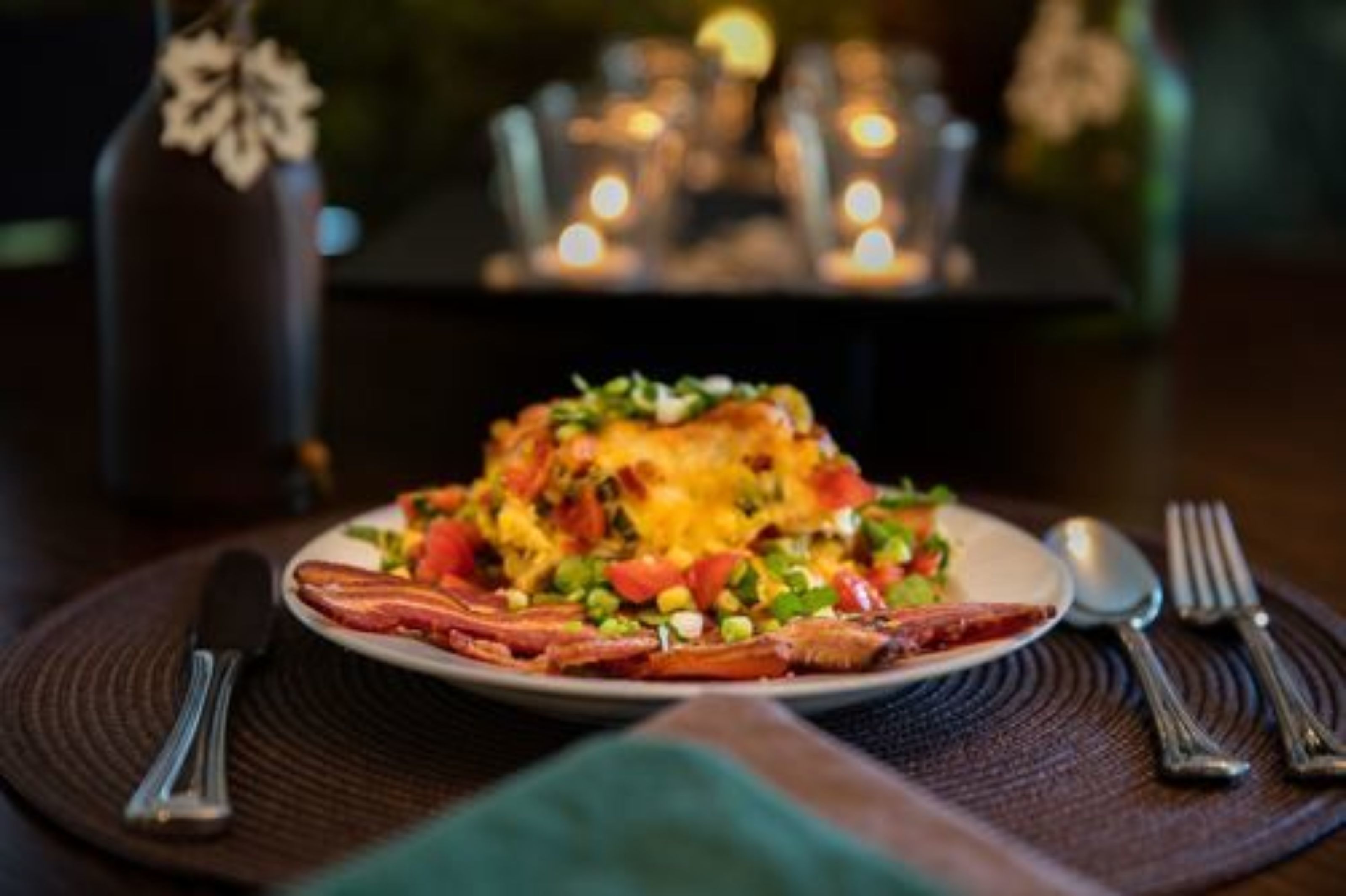 A plate of food with bacon, cheese, and vegetables on a table with candles in the background.