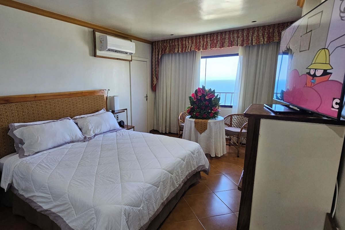 A hotel room with a bed, a desk with a chair, an air conditioner, a TV, and a window with curtains.
