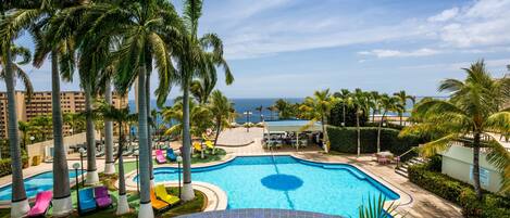 A resort pool area with palm trees, lounge chairs, and a view of the ocean.