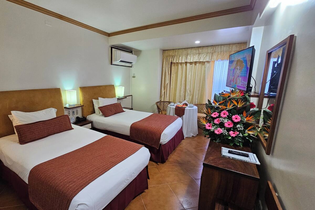 A hotel room with two beds, a desk, a chair, a flower arrangement, and a painting on the wall.