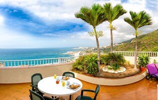 Terraza con mesa lista para comer, con vista al mar, palmeras y un paisaje urbano distante.