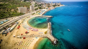 An aerial view of a coastal city with a beach, clear blue waters, and a mix of residential and commercial buildings. - Hotel Ole Caribe (Macuto)