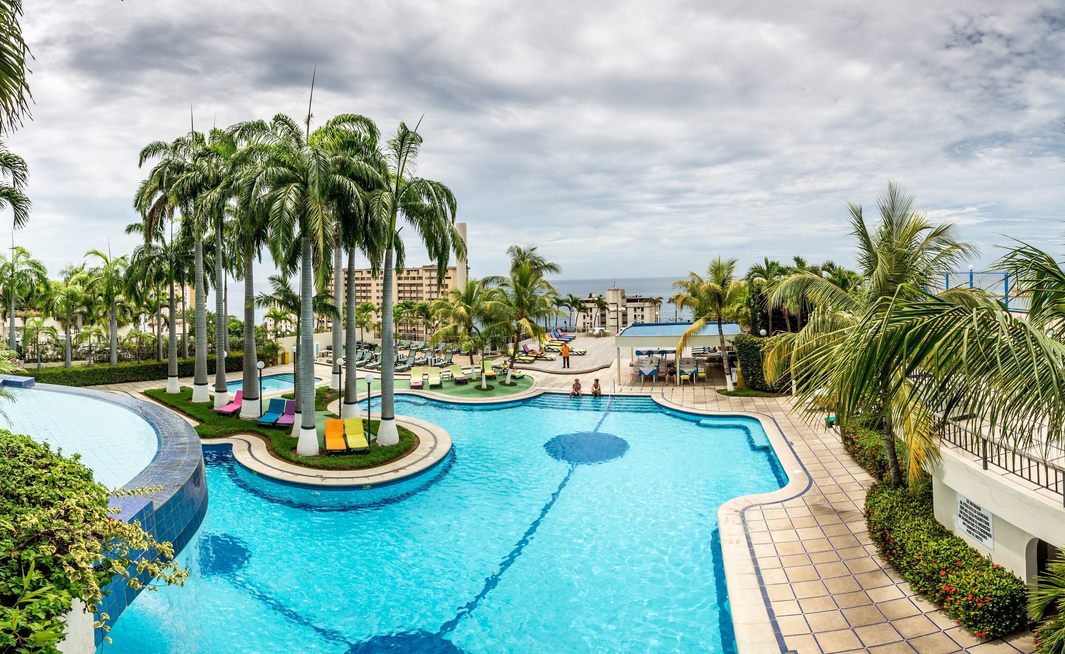 A large outdoor swimming pool surrounded by palm trees and lounge chairs, with a view of the ocean in the background.