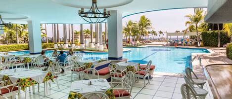 A poolside area with white wicker chairs and tables, a pool, and palm trees in the background.