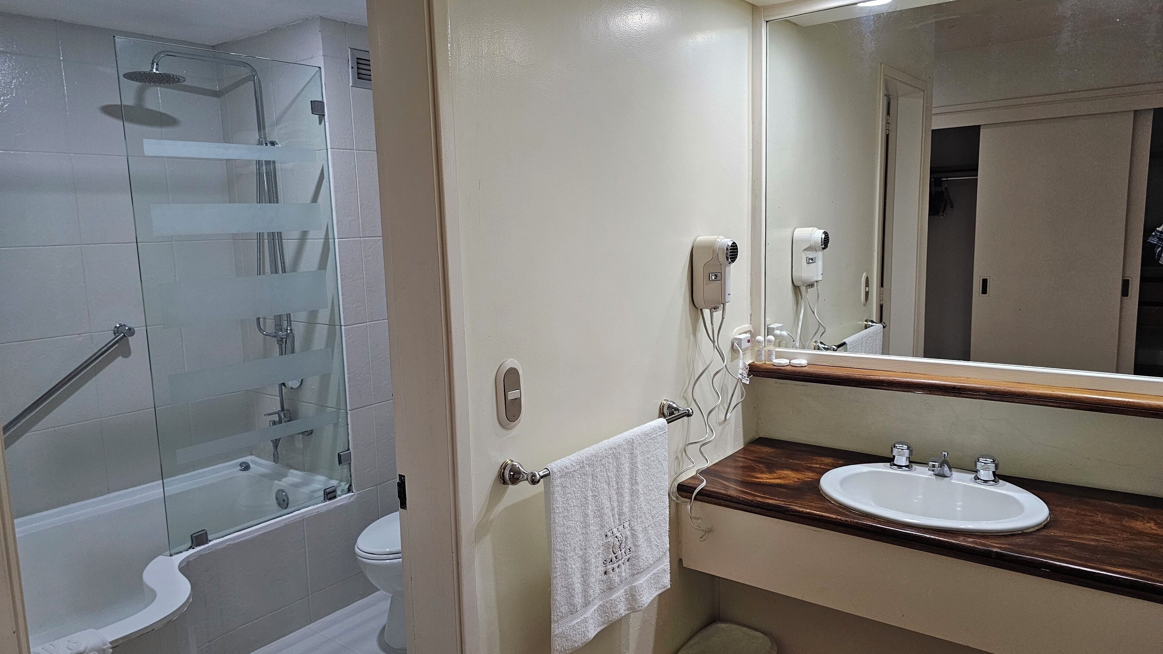 Un baño con ducha de mampara de vidrio, inodoro blanco, lavabo con encimera de madera y espejo con una toalla colgada en la pared.