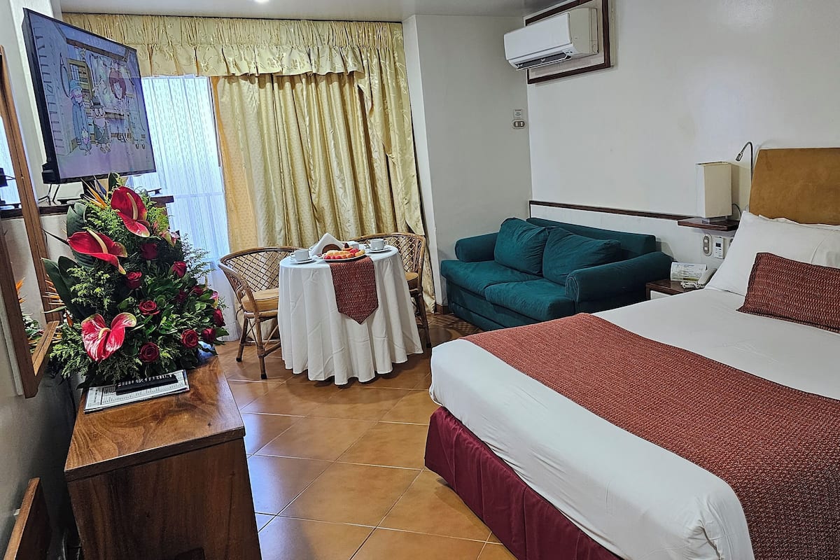 A hotel room with a bed, a green sofa, a small table with a white tablecloth, a decorative plant, and a television on the wall.