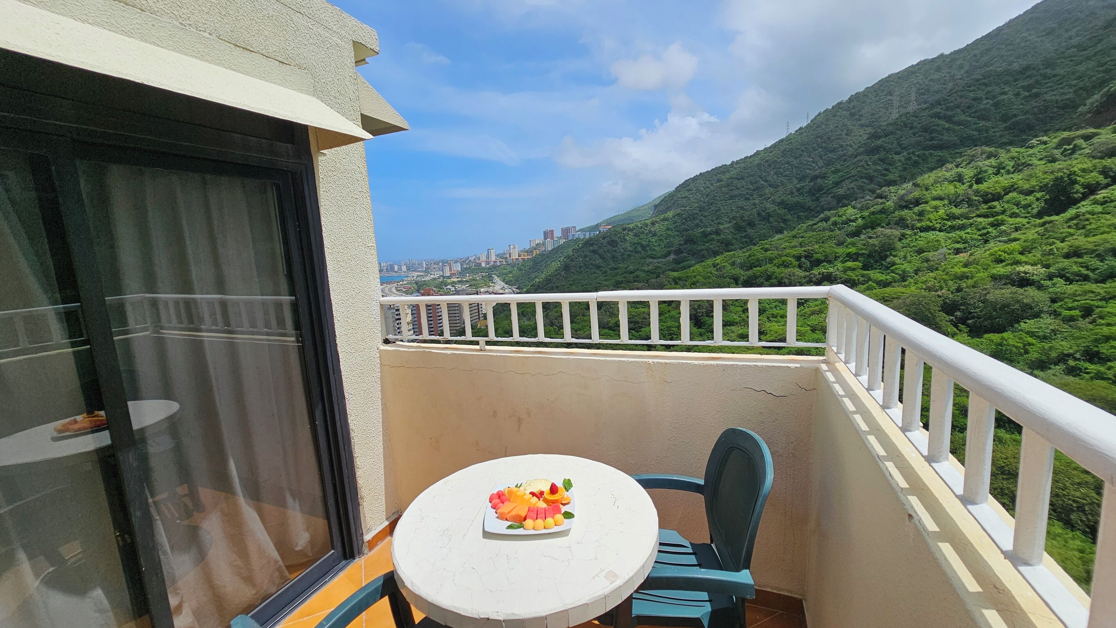 A balcony with a table and chairs, overlooking a cityscape and lush green hills.