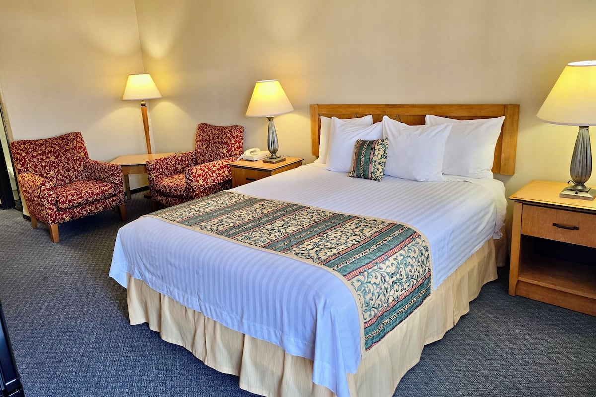 A hotel room with a large bed, two armchairs, a nightstand with a lamp, and a bedside table with a lamp.