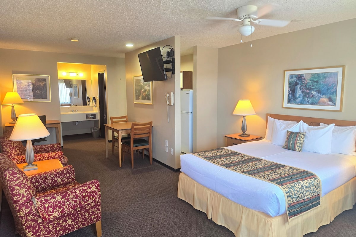 A hotel room with a bed, two chairs, a desk, a television, and a ceiling fan.