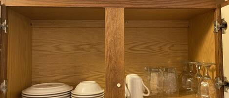 Un mueble de madera con dos estantes, uno con platos apilados y el otro con una jarra blanca y varios vasos de vidrio.