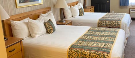 Habitación de hotel con dos camas, cada una con cabecero de madera, ropa de cama blanca y almohadas decorativas. Hay dos mesitas de noche con lámparas, un aparador de madera y una puerta con panel oscuro.