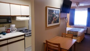 Una kitchenette compacta con estufa, microondas y cafetera. Un cuadro enmarcado cuelga en la pared, y se ve una cama con colcha estampada al fondo.