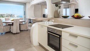 A modern kitchen with a central island, built-in oven, and a dining area with striped chairs and a table. - Villa Melmastia (Mykonos)