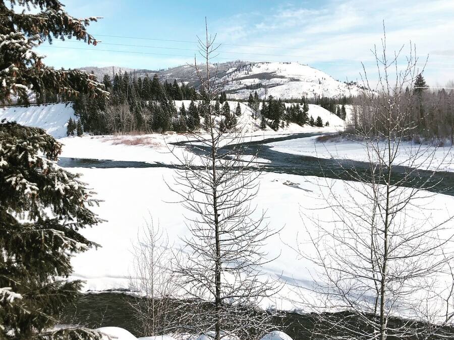 Un paisaje nevado con un río, árboles y montañas a lo lejos.