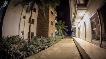 Un pasillo nocturno con una palmera y arbustos, que conduce a un edificio de varios pisos y balcones.
