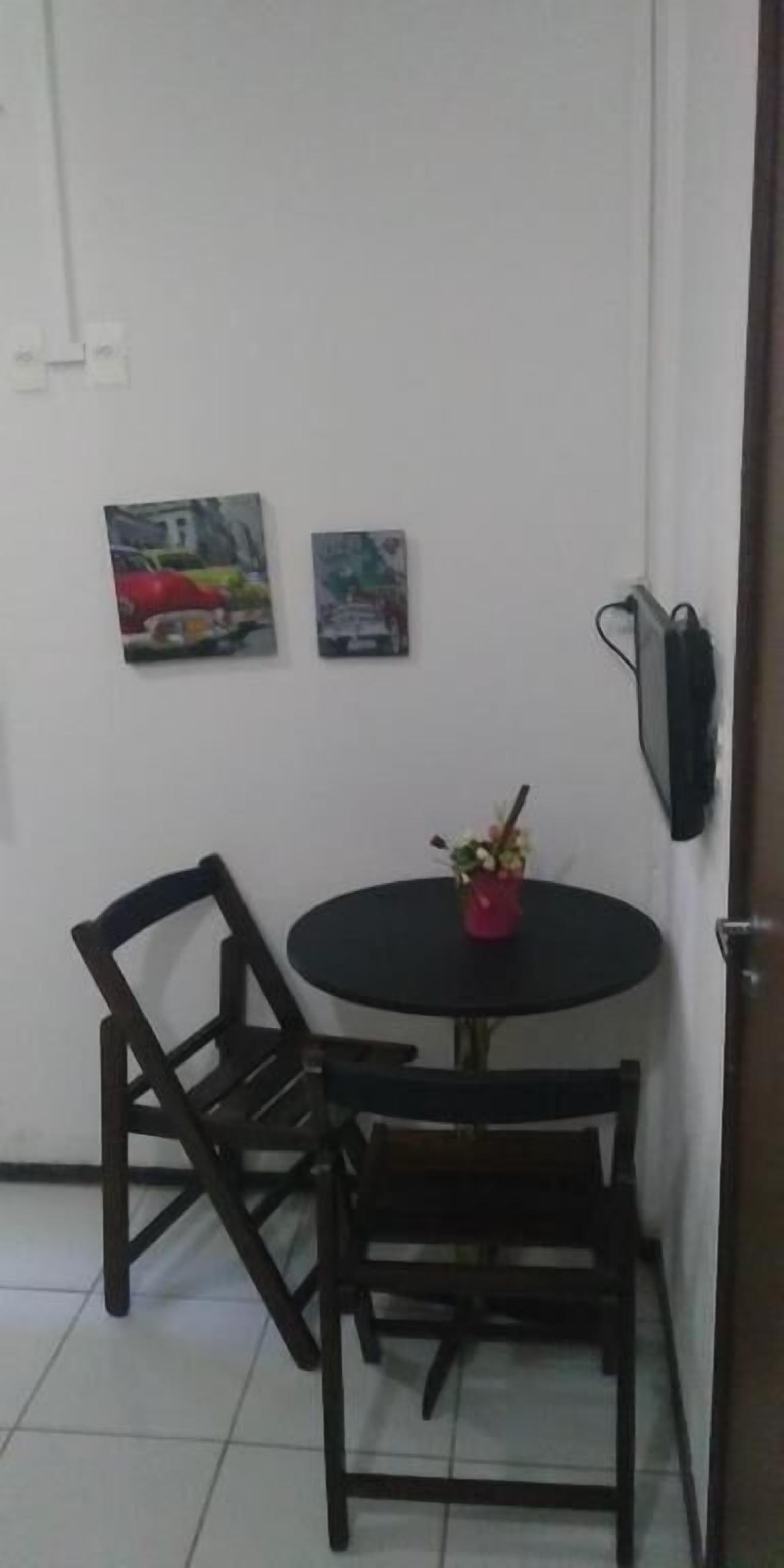 Un pequeño comedor con una mesa redonda y dos sillas, un pequeño jarrón rojo con flores, y dos cuadros en la pared.