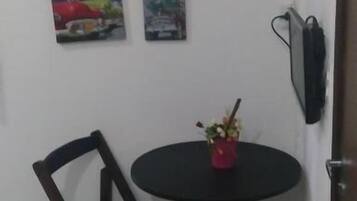 Un pequeño comedor con una mesa redonda y dos sillas, un pequeño jarrón rojo con flores, y dos cuadros en la pared.