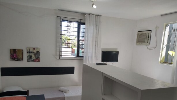 A dormitory room with a bed, a desk, a chair, a TV, and a window with curtains. - Studio Boa Viagem (Recife)