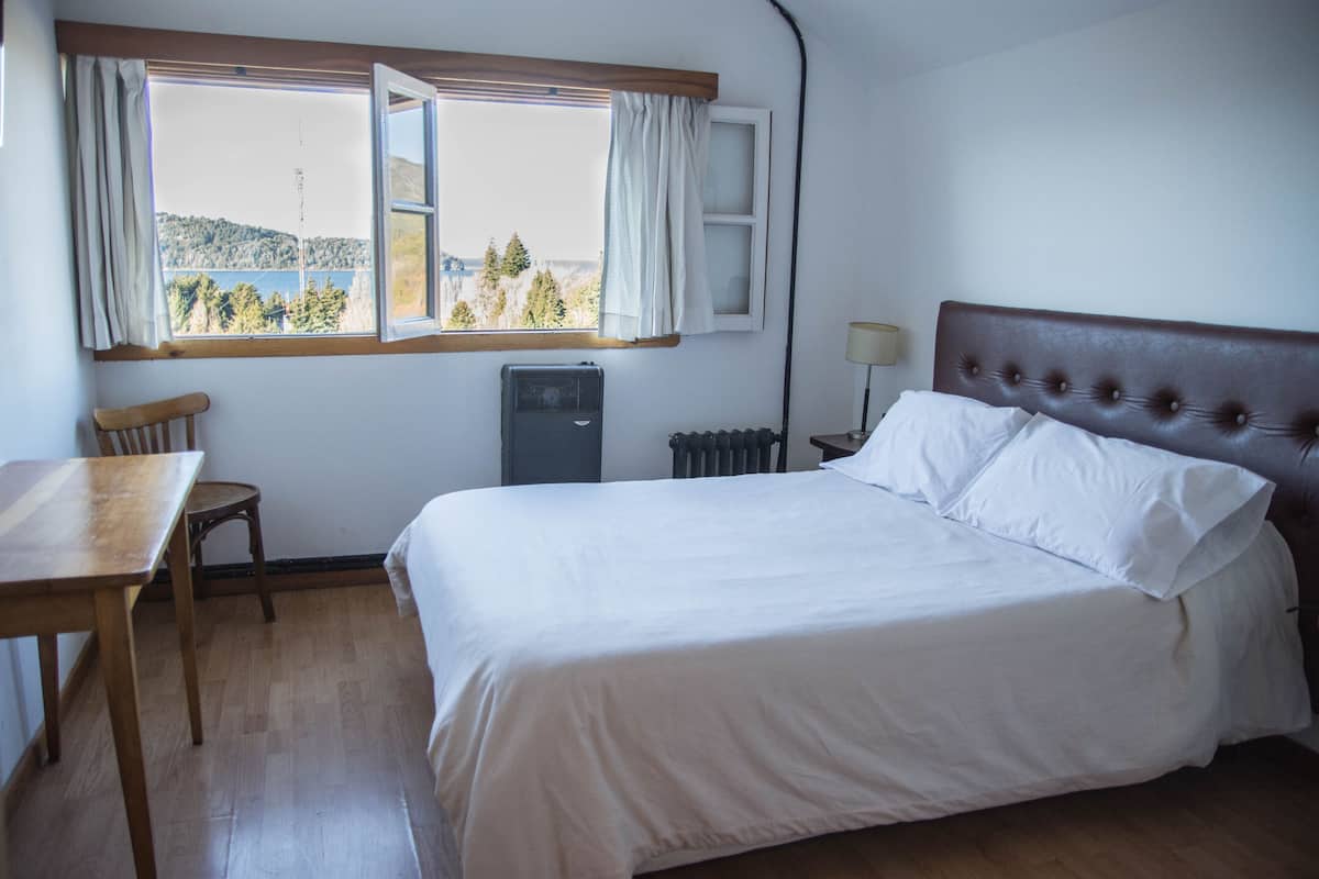 Une chambre à coucher avec un lit, une table, une chaise, un radiateur et une fenêtre avec des rideaux.