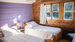 A room with two wooden beds, a window with curtains, a radiator, and a chair. - Hosteria Pajaro Azul (San Carlos de Bariloche)