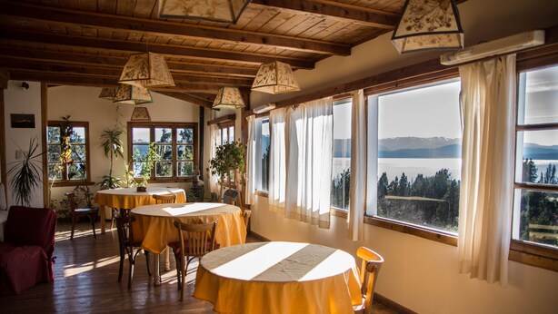 A dining area with wooden tables and chairs, large windows with curtains, and hanging lamps. - Hosteria Pajaro Azul (San Carlos de Bariloche)