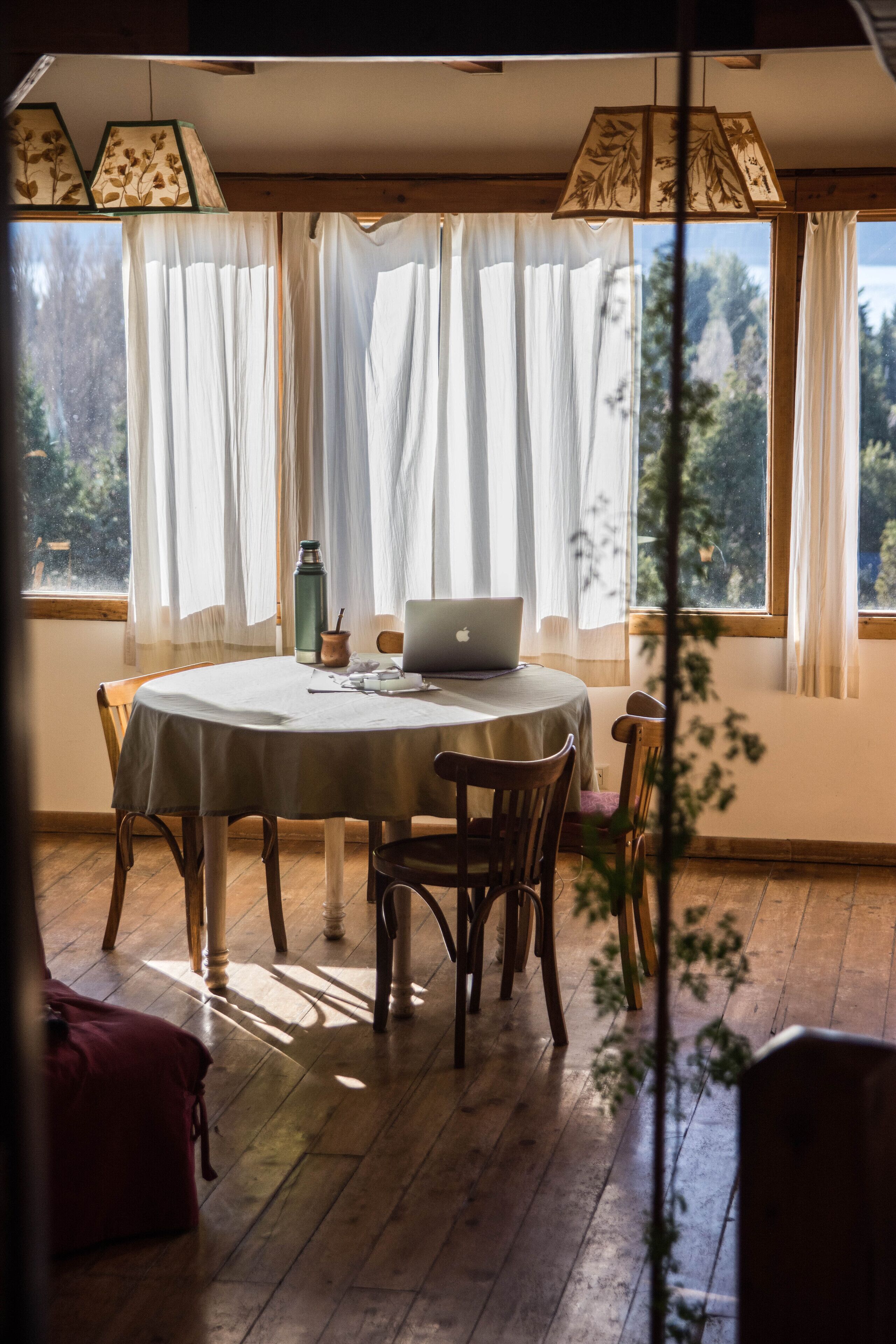 A dining area with a round table, chairs, a laptop, and a view through large windows.