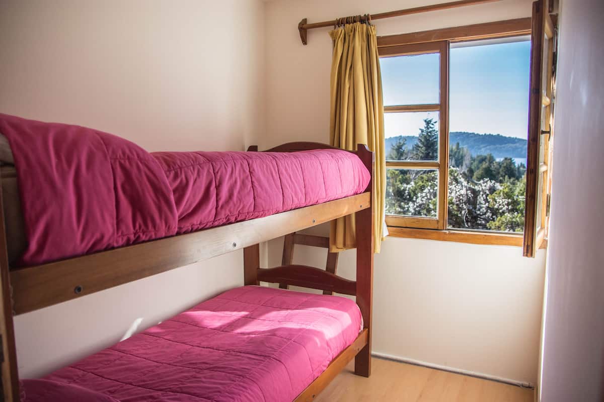 Un lit superposé avec une literie rose, dans une chambre dont la fenêtre donne sur une zone boisée.