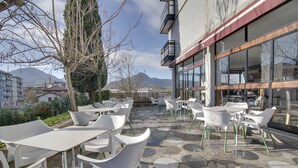 An outdoor seating area with white plastic chairs and tables, a stone-tiled floor, and a building with large windows in the background. - Hotel San Blas (Abadino)