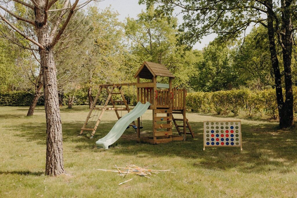 Un patio de juegos con un tobogán de madera y pared de escalar, rodeado de árboles y césped.