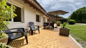 An outdoor patio with a tiled floor, a wooden table with chairs, a black plastic chair, and a wooden planter.
