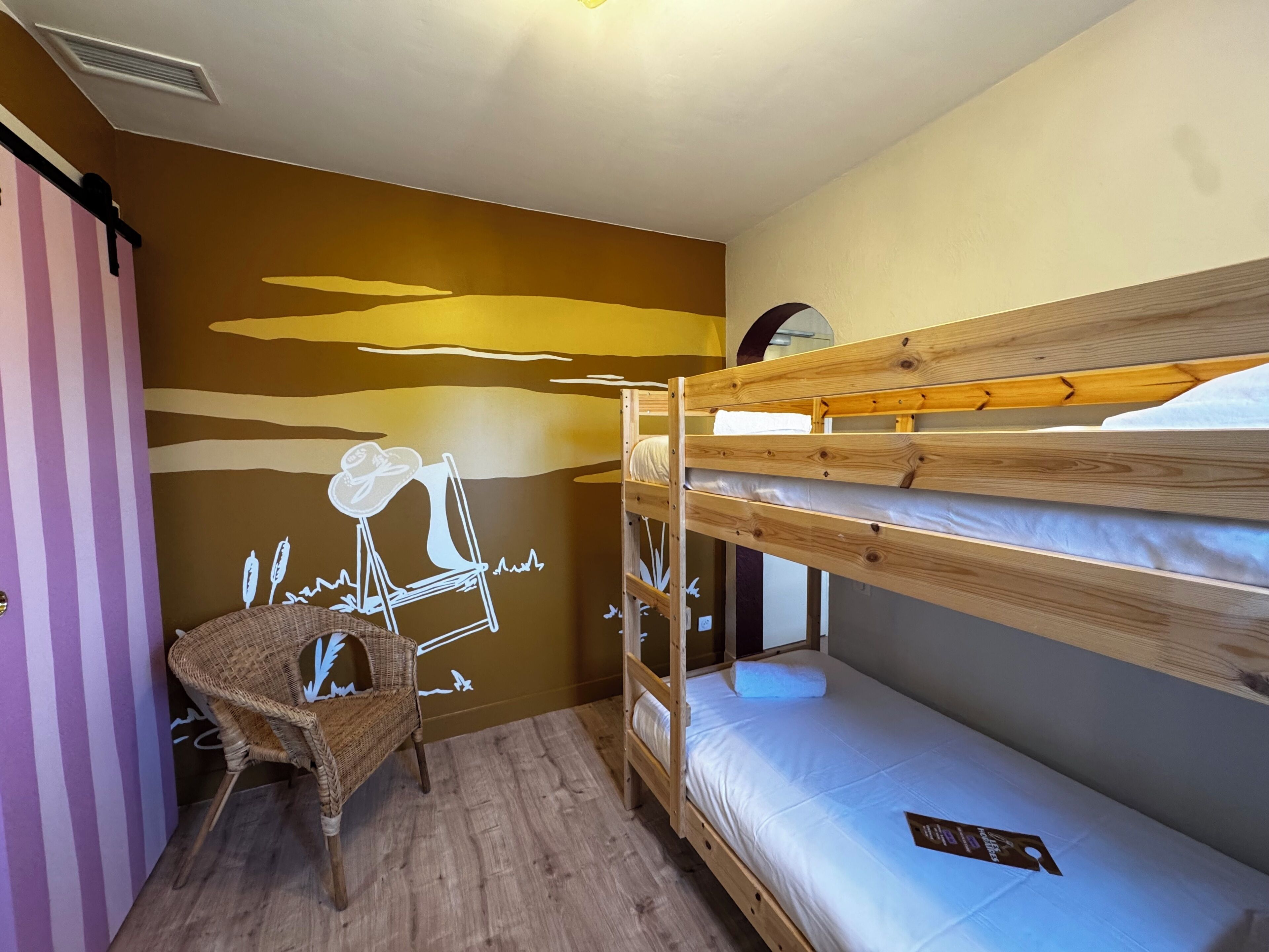 Dormitorio con literas, suelo de madera, una silla de mimbre y un mural en la pared.