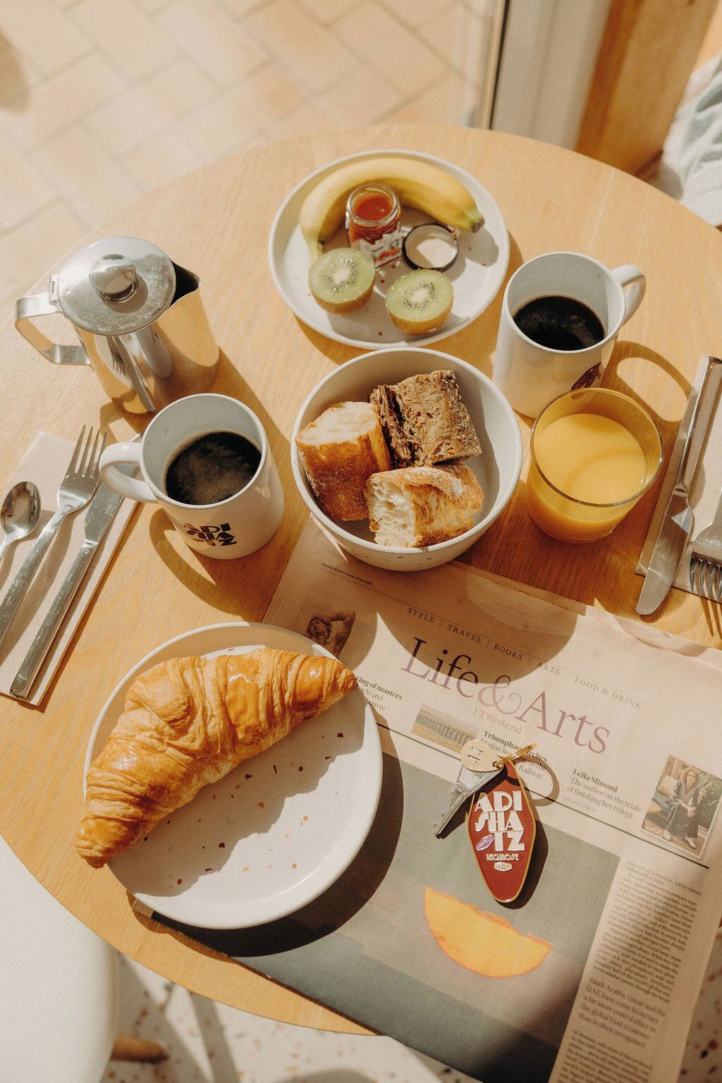 Desayuno con croissants, tostadas, plátano, kiwi, mermelada, café y un periódico.
