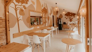 A restaurant with wooden tables and white chairs, a large mirror, and a wall mural of trees and leaves.