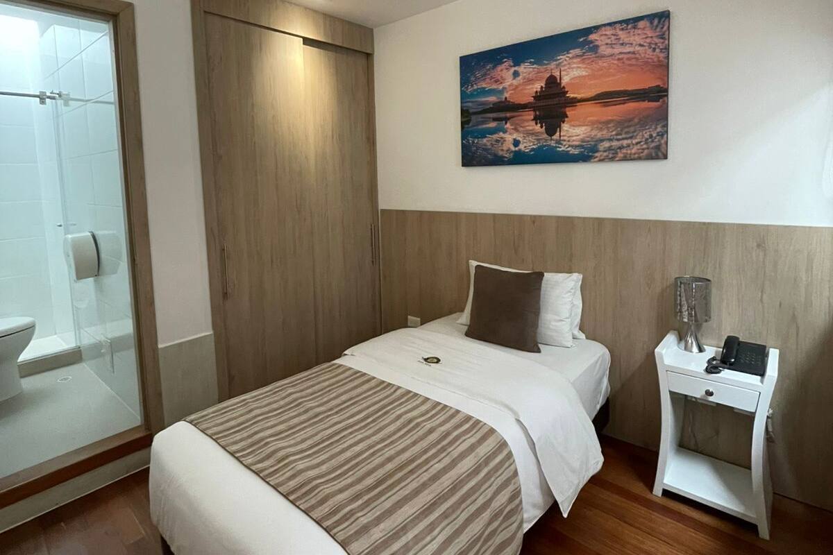 Una habitación de hotel moderna con una cama grande, una mesita de noche con lámpara, un cuadro en la pared y una cabina de ducha de vidrio.