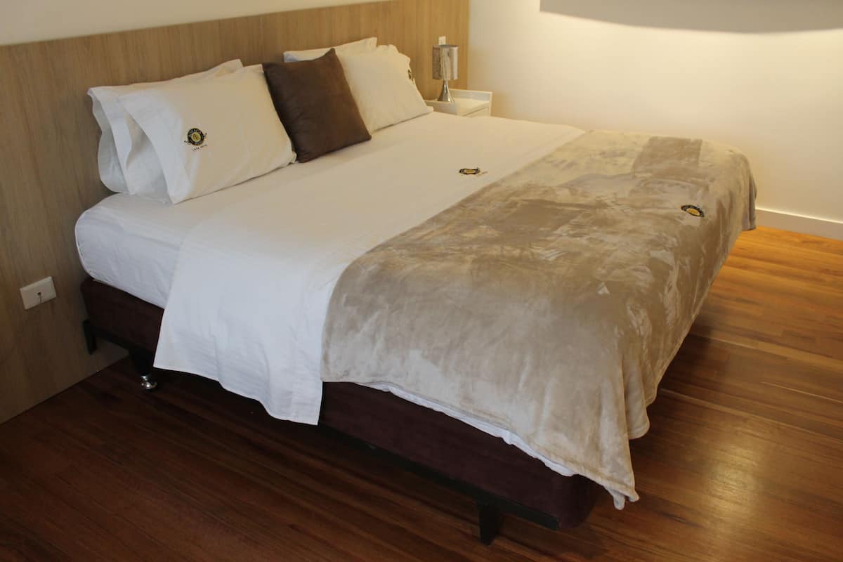 Una cama bien tendida con cabecera de madera, una mesita de noche con lámpara y suelo de madera.