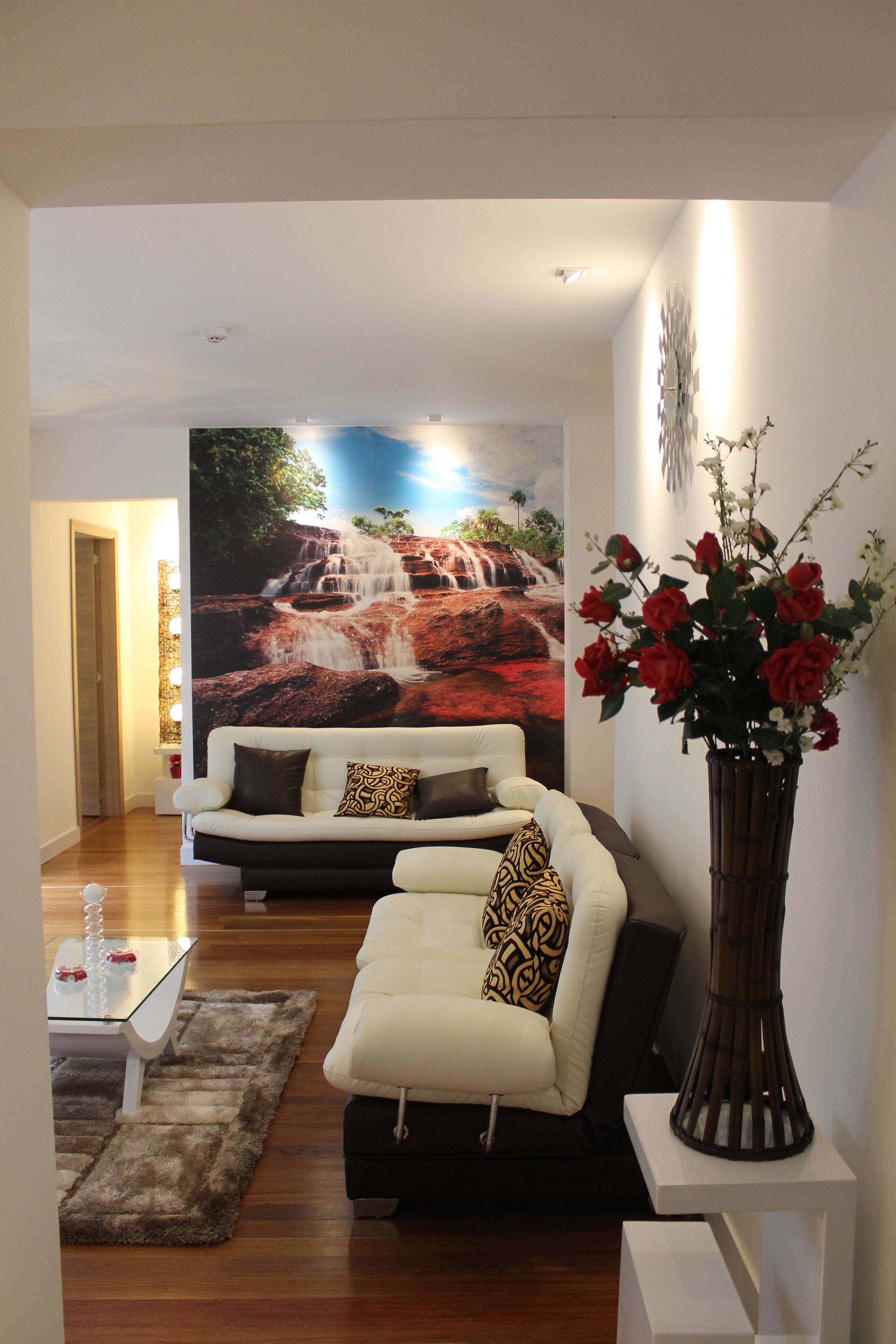 A modern living room with a white sofa, a glass coffee table, and a large waterfall mural on the wall.