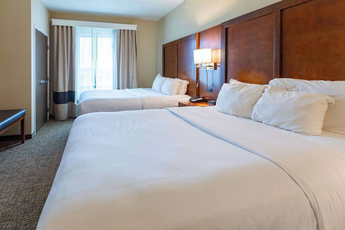 Une chambre d’hôtel avec deux lits, une table de chevet avec une lampe, une fenêtre avec des rideaux et une porte.