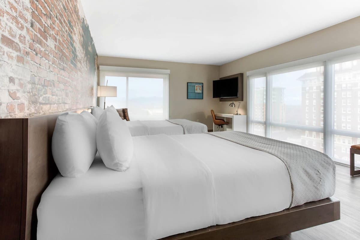 A hotel room with a large bed, a desk, a chair, a TV, and large windows.