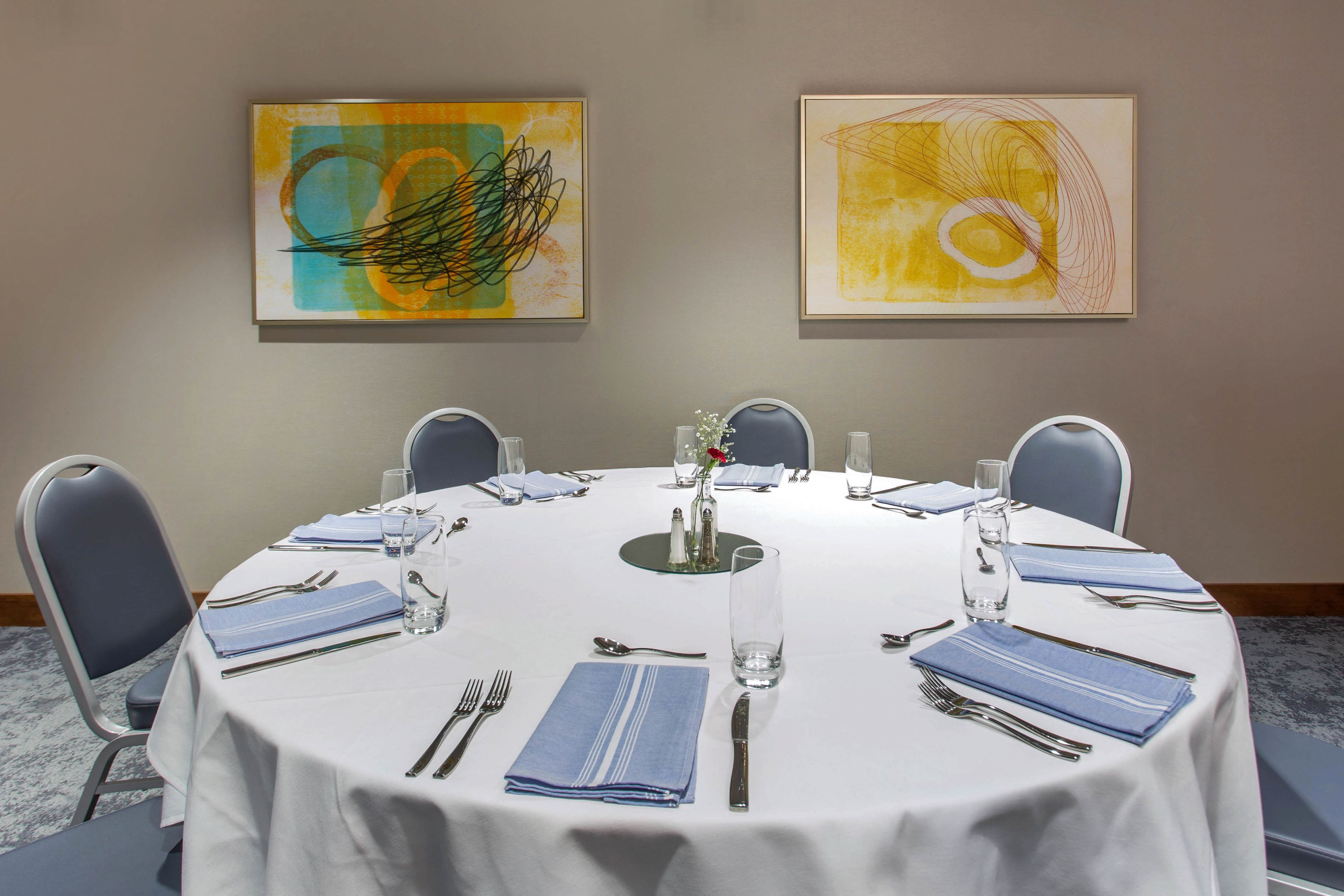 A conference room with a round table set for six, blue chairs, and two abstract paintings on the wall.