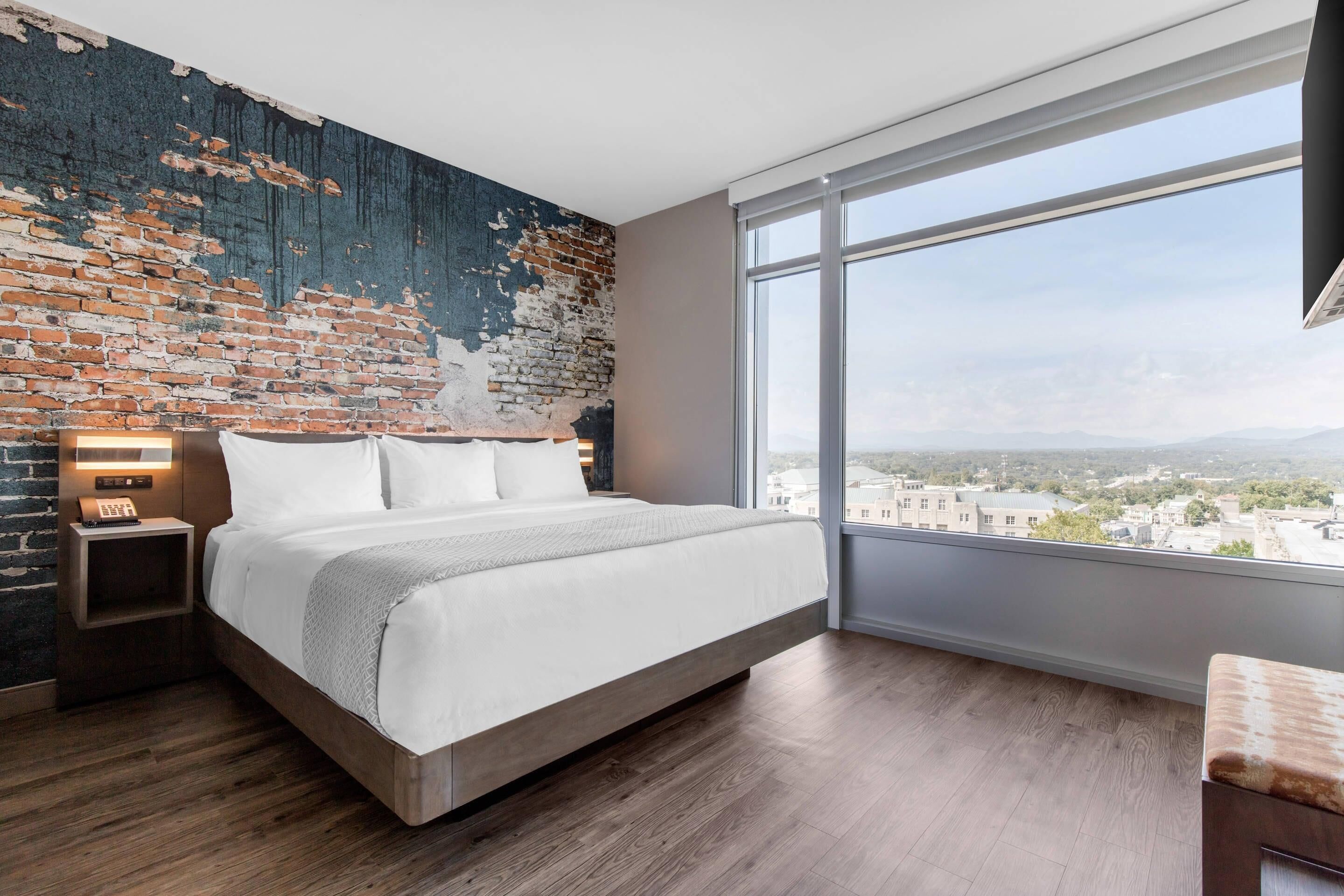 Une chambre moderne avec un grand lit, un mur en briques, une table de chevet et une vue sur la ville par une grande fenêtre.
