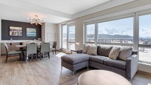A modern living room with a gray sectional sofa, a dining area with a table and chairs, and large windows offering a view of a waterfront and distant hills. - Cambria Hotel Downtown Asheville (Asheville)