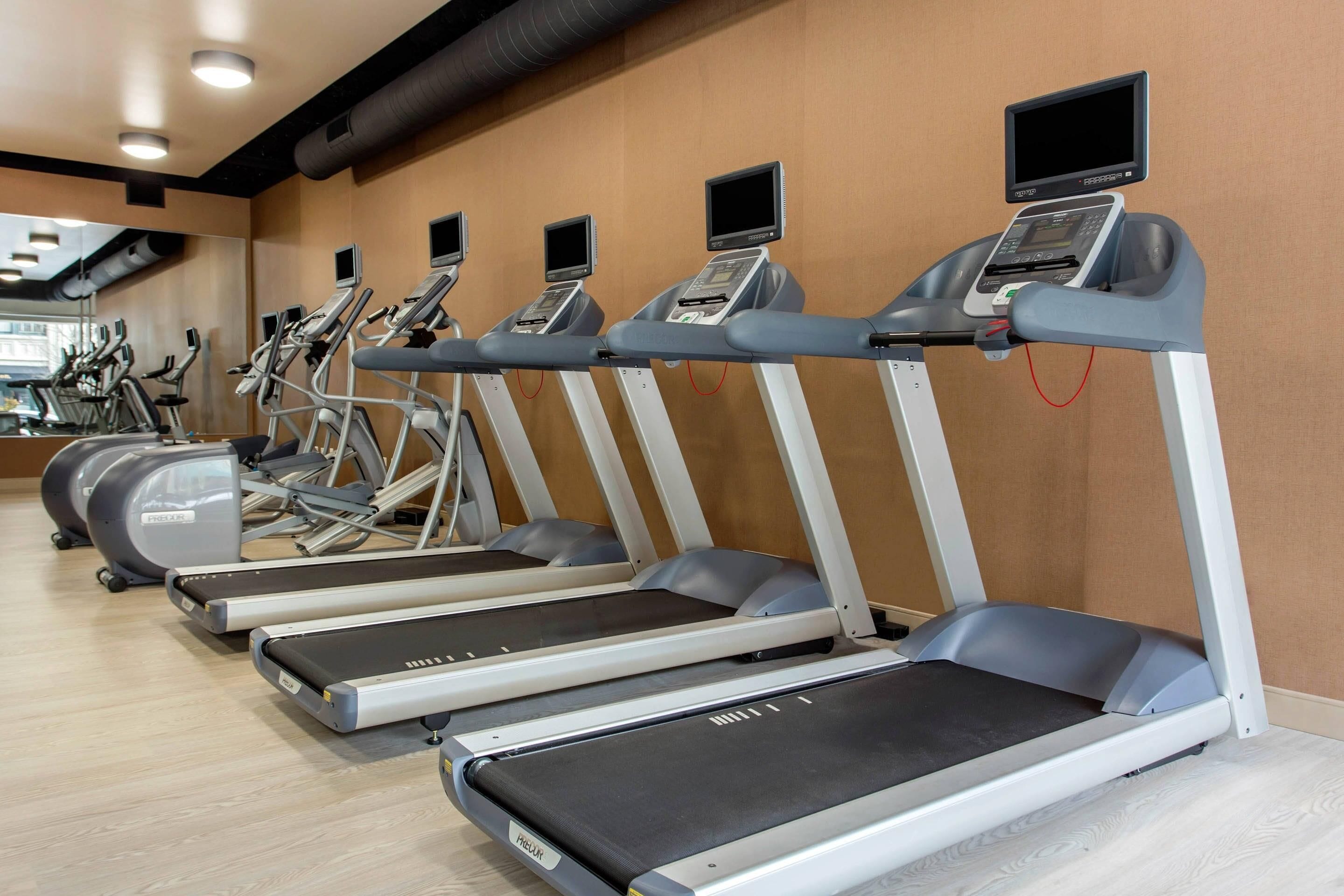A row of treadmills with digital screens, a mirrored wall, and exercise bikes in a gym.